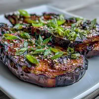 Golden-brown miso glazed eggplant halves with caramelized edges, garnished with toasted sesame seeds and fresh green onions on a rustic wooden board.
